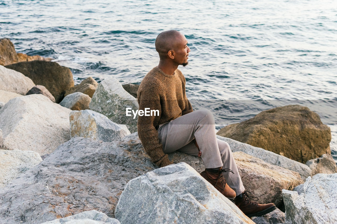 Side view full body of calm african american male looking into distance with pensive face while sitting on rocky coast near sea