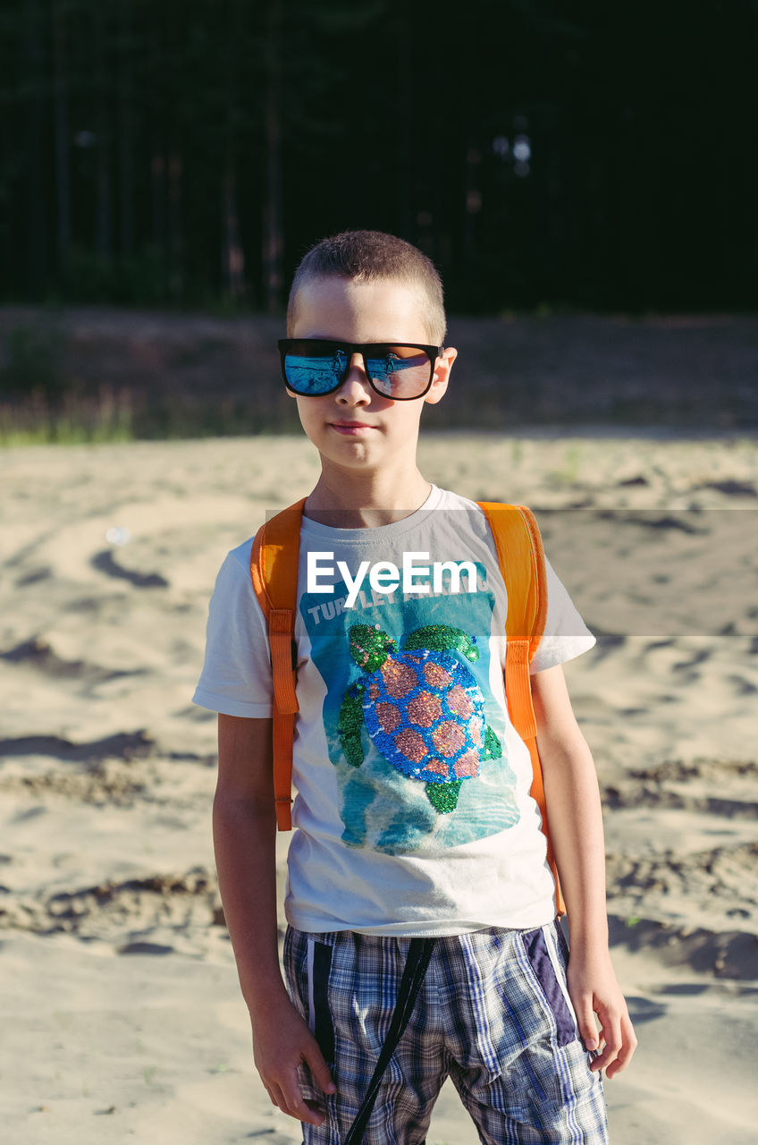 A seven-year-old boy in shorts, a t-shirt and sunglasses stands on a sandy glade