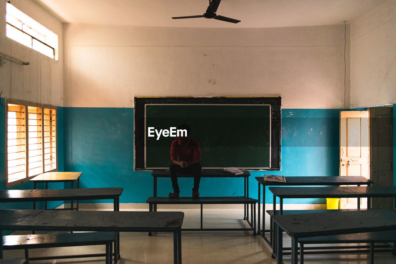 Teenage boy sitting on table against blackboard in school