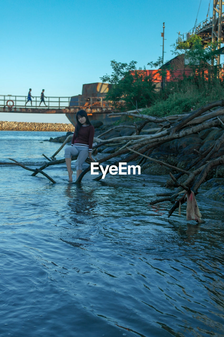 Teenage girl sitting on branch in river against clear sky