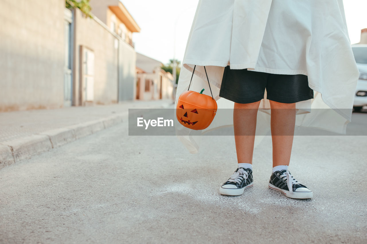 Cropped unrecognizable kid in white ghost costume and with plastic jack o lantern standing in city on halloween in autumn