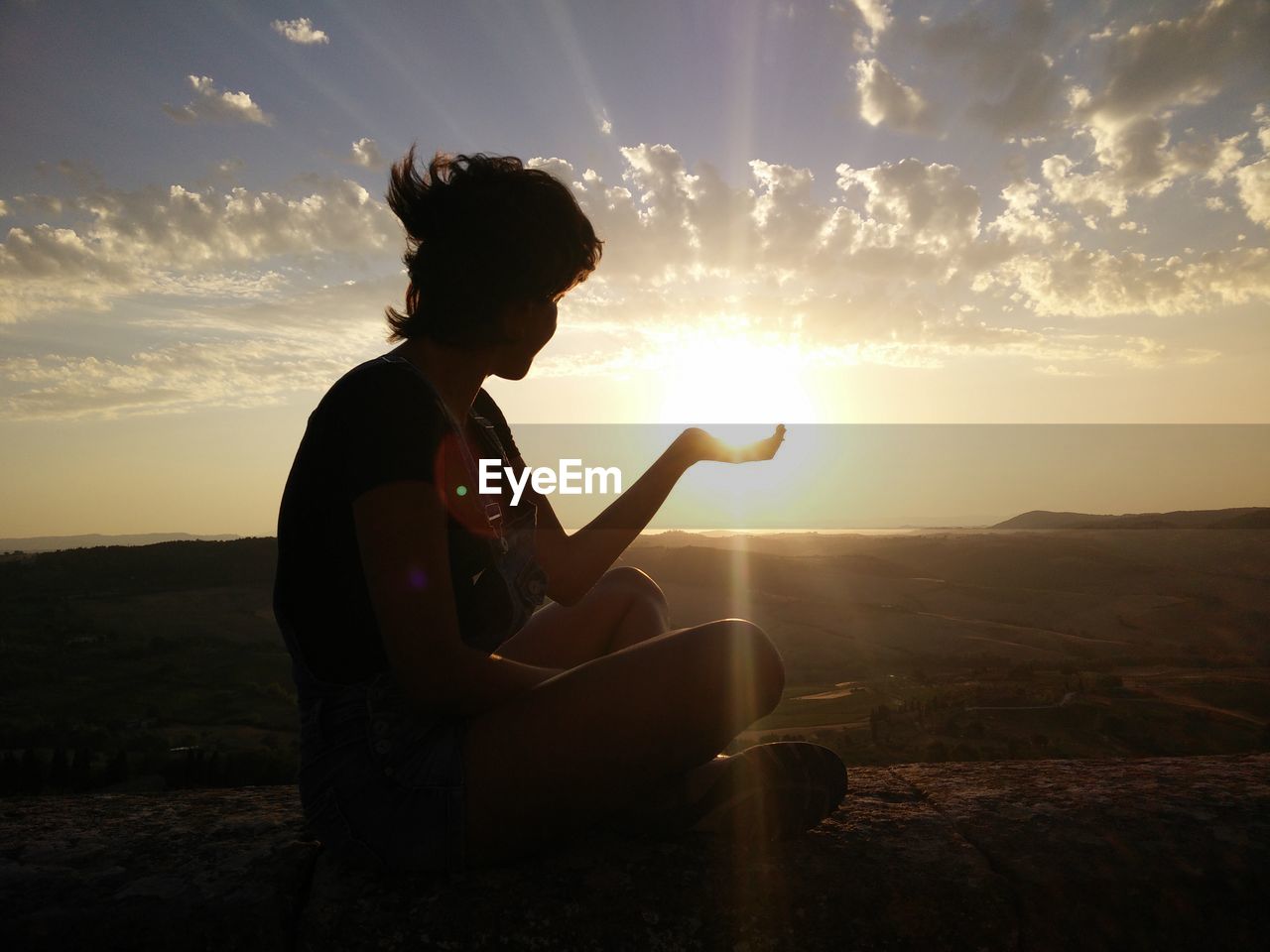 Silhouette woman gesturing while sitting on retaining wall against sky during sunset