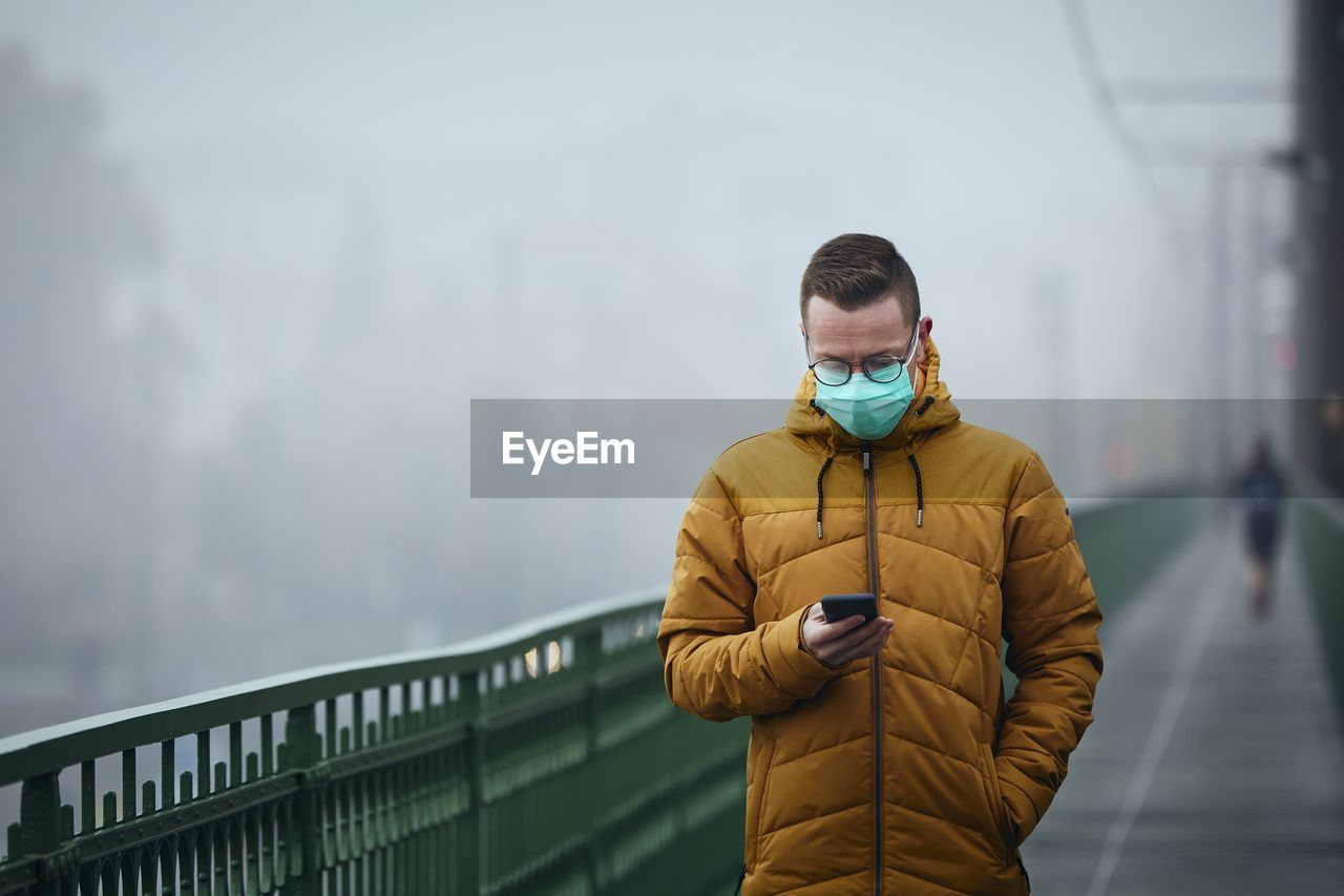 Man wearing mask using smart phone standing by railing during winter