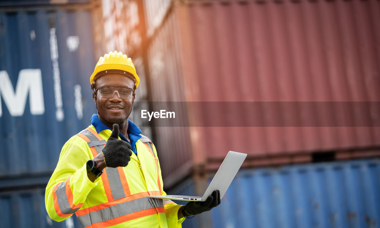 Portrait of worker holding laptop showing thumbs up while standing at dock