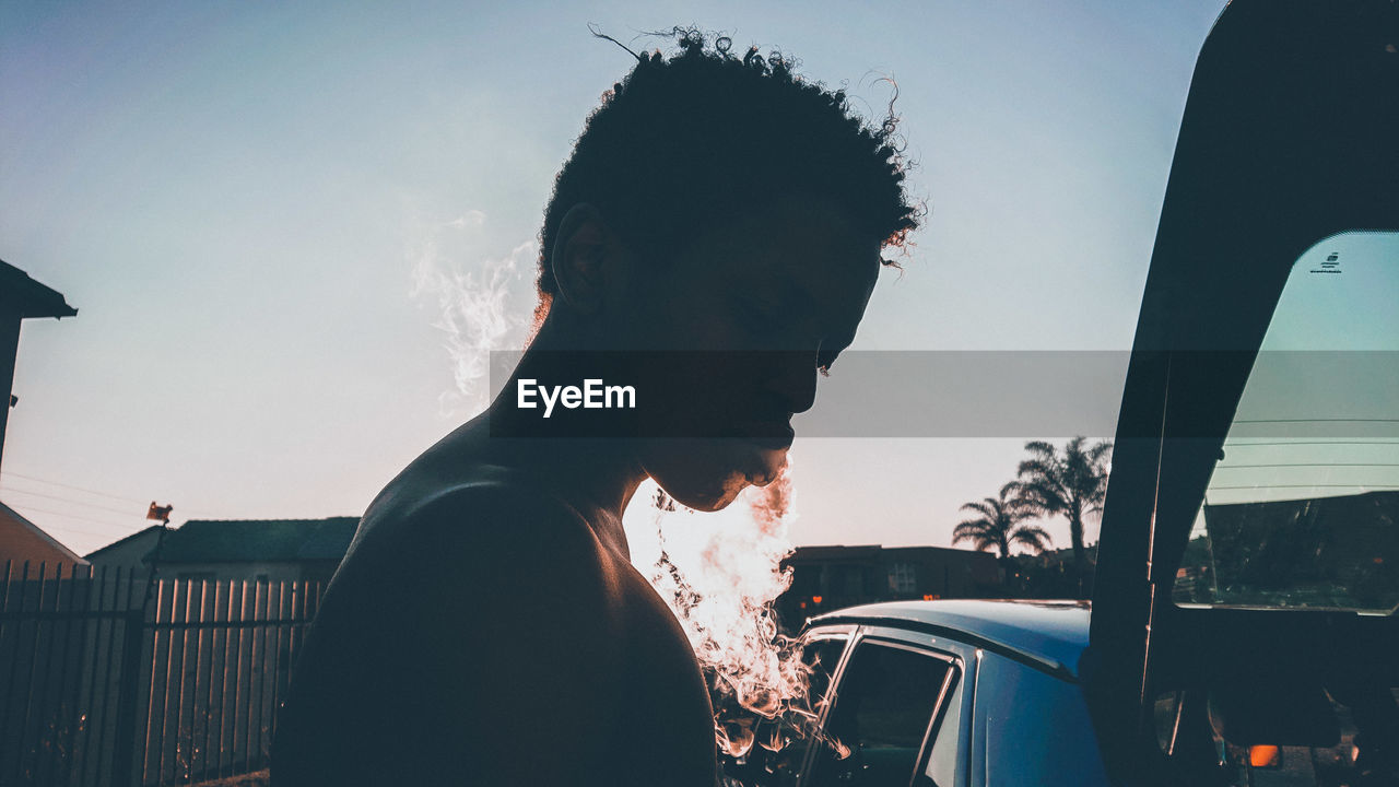 Side view of young man smoking while standing against clear sky during sunset