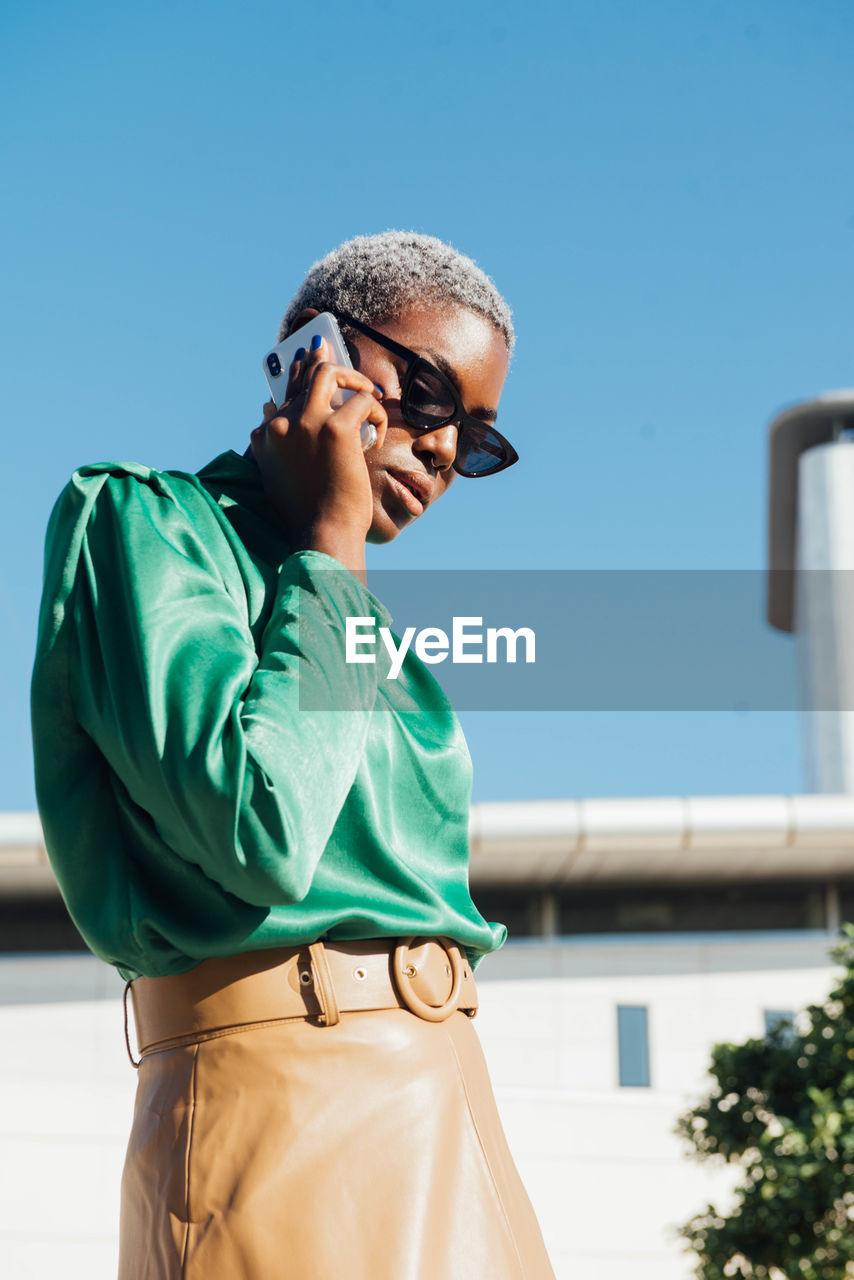 Confident young african american businesswoman with short dyed hair in stylish clothes and sunglasses talking on mobile phone while standing on street against cloudless blue sky