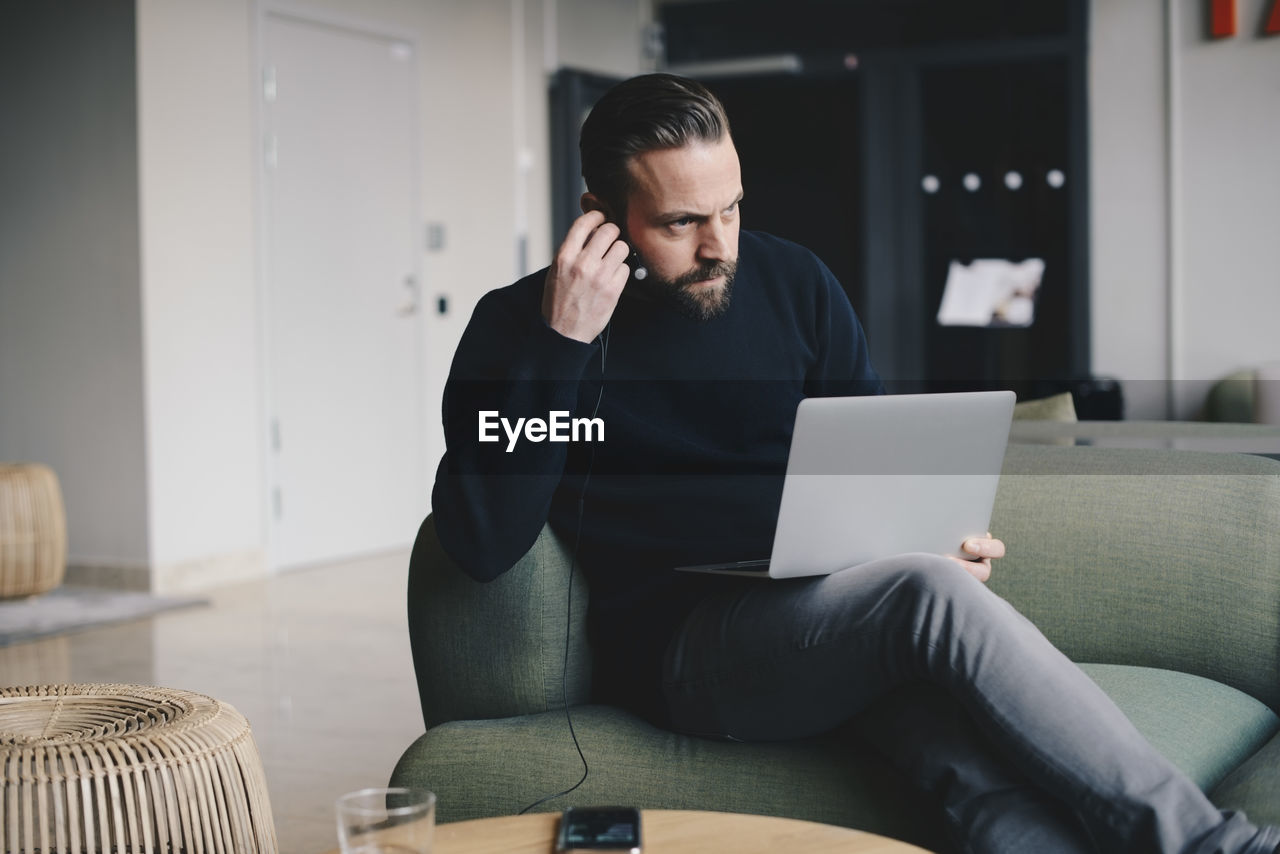 Businessman wearing in-ear headphones while sitting with laptop on sofa at office lobby