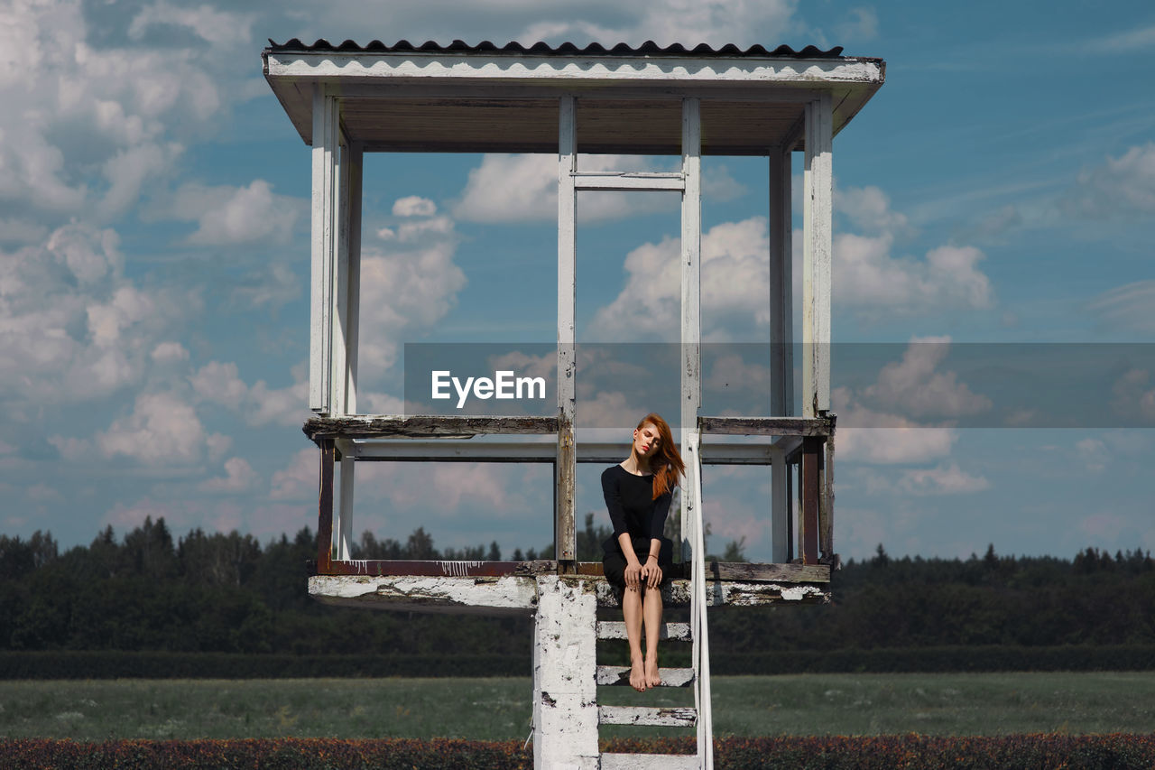 Woman sitting at lookout tower on field against sky