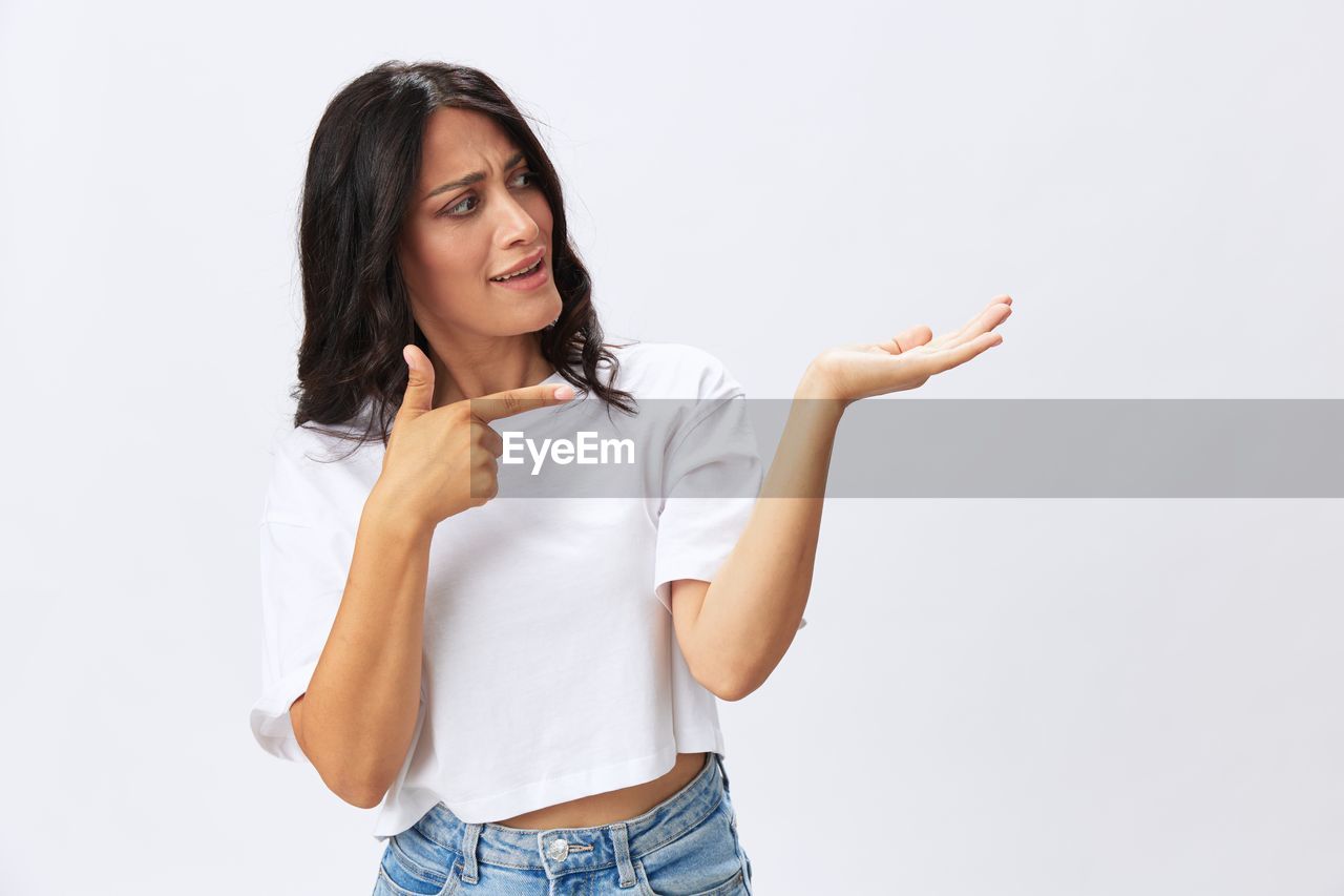 one person, women, adult, studio shot, casual clothing, indoors, portrait, young adult, white background, emotion, smiling, sleeve, clothing, photo shoot, happiness, copy space, jeans, standing, female, long hair, front view, gesturing, cut out, looking, positive emotion, looking at camera, person, hairstyle, hand, cheerful, finger, lifestyles, relaxation, brown hair, communication, waist up, t-shirt, arm, limb