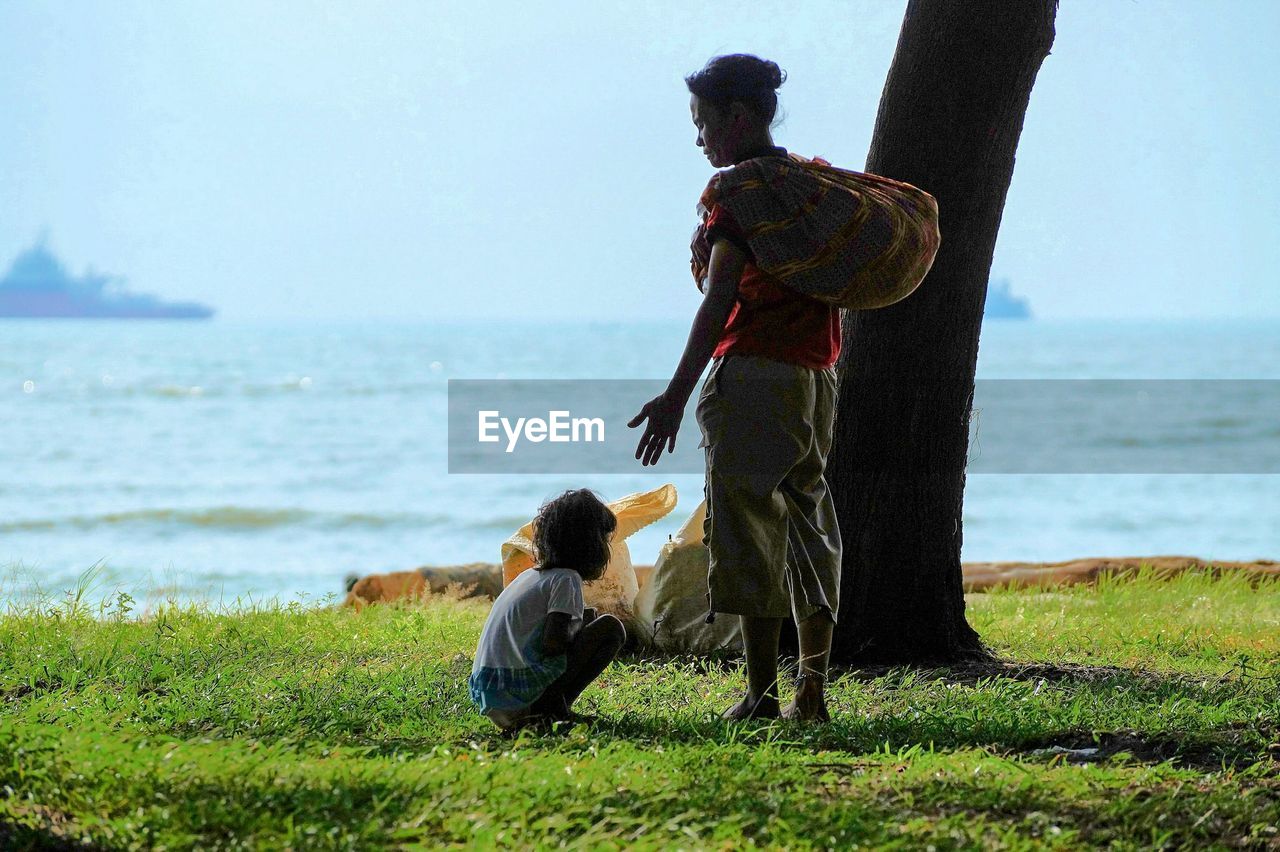 Rear view of woman standing by daughter crouching on grassy field against sea