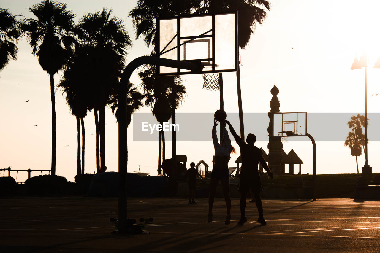 Silhouette friends playing basketball against sky during sunset