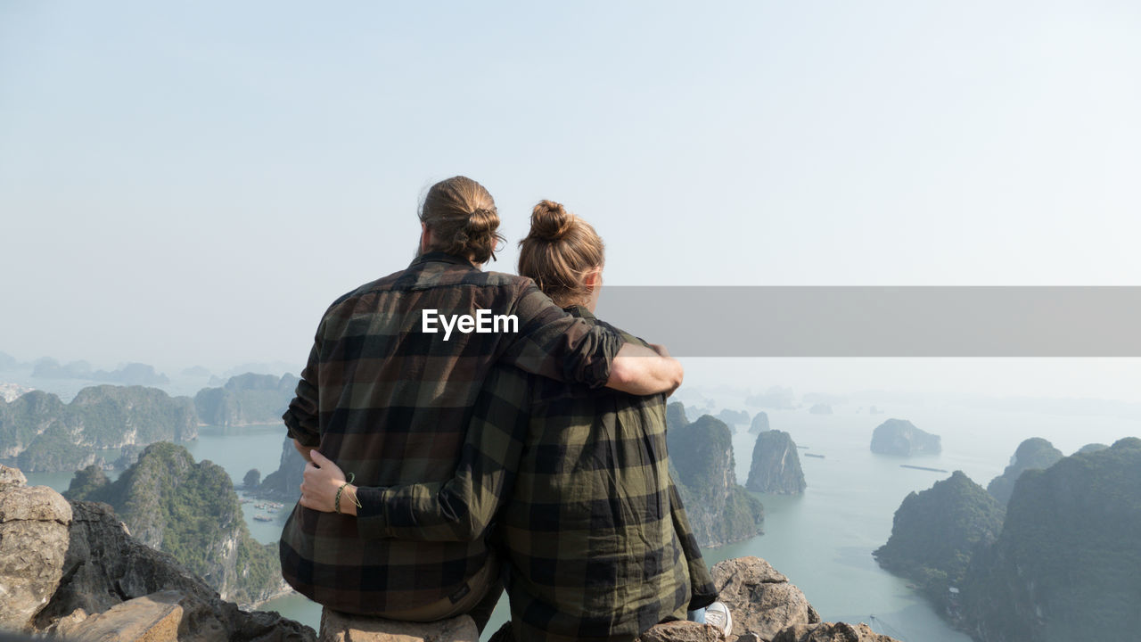 Rear view of couple sitting on rocks against sky