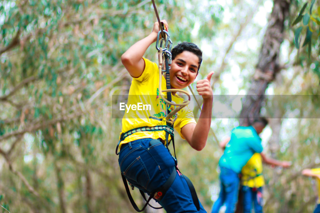 Portrait of smiling teenage boy zip lining with man in background at forest