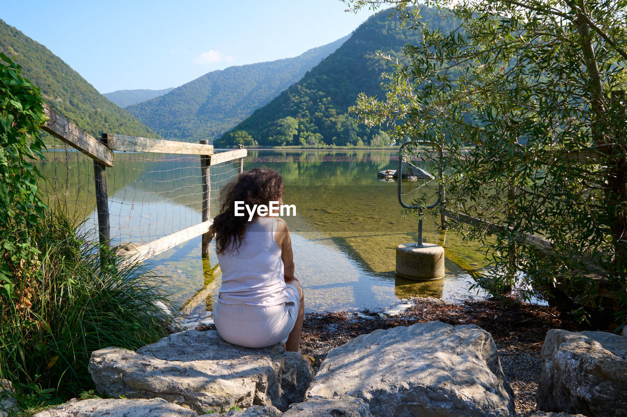 rear view of young woman sitting on rock by lake