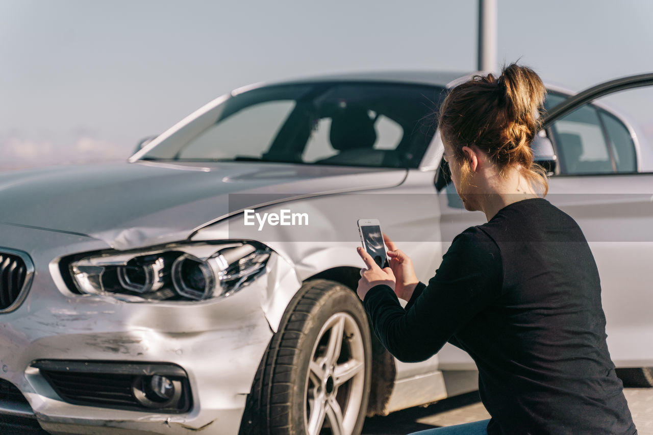 Side view of unrecognizable young female driver in casual clothes taking photo of car damages on smartphone after accident on road on sunny day