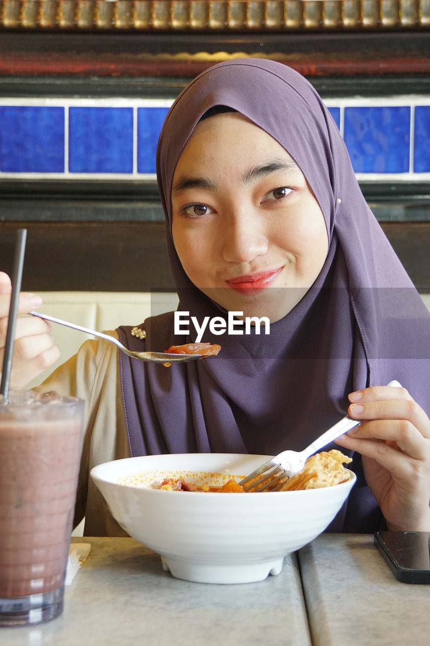 Portrait of young woman wearing hijab while eating food at restaurant