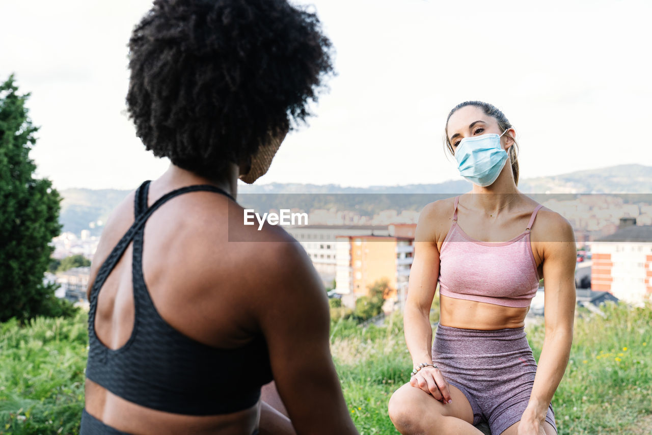 Young fit female friends in activewear and protective masks sitting on wooden fence and chatting while having break during outdoor training in summer day