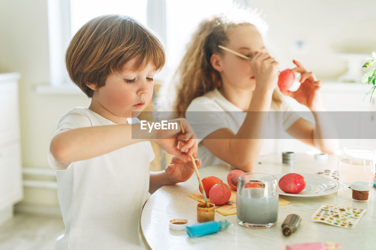 Two siblings brother and sister toddler boy tween girl painting easter eggs on kitchen at home 