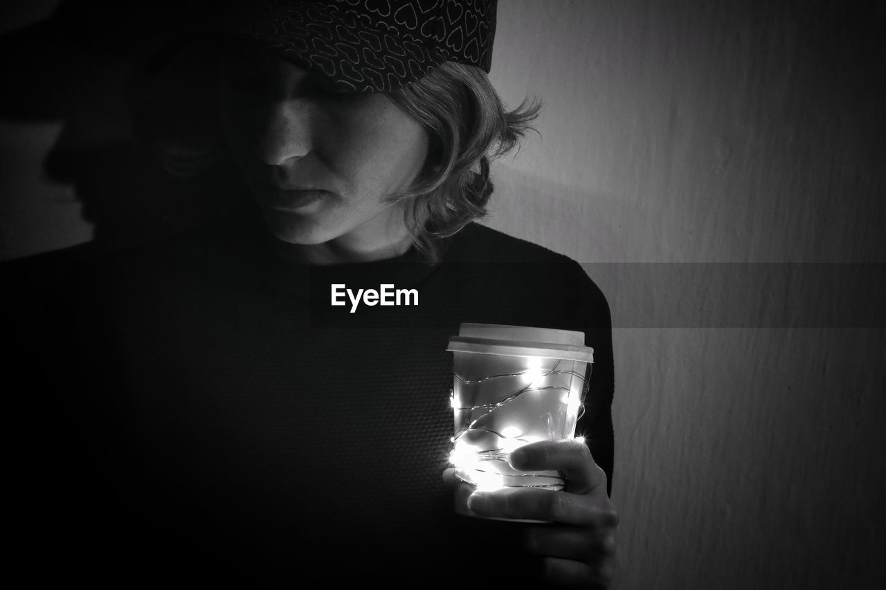 Close-up of mid adult woman holding illuminated string lights with coffee cup against wall in darkroom