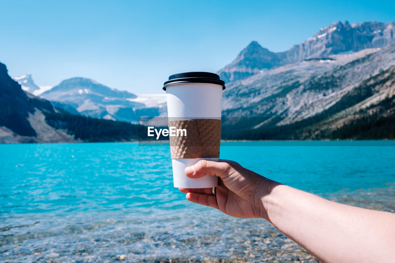 Cropped image of hand holding drink against lake and mountains