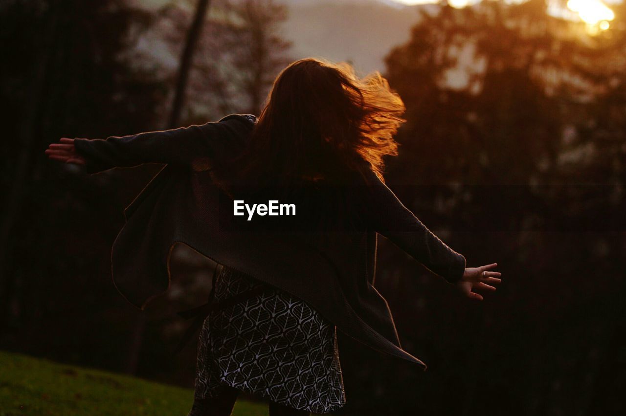 Rear view of cheerful woman with arms outstretched dancing at park during sunset