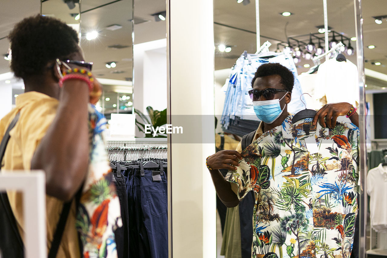 Young african american male in trendy outfit and medical mask standing in front of mirror in modern boutique and picking goods in shop during coronavirus