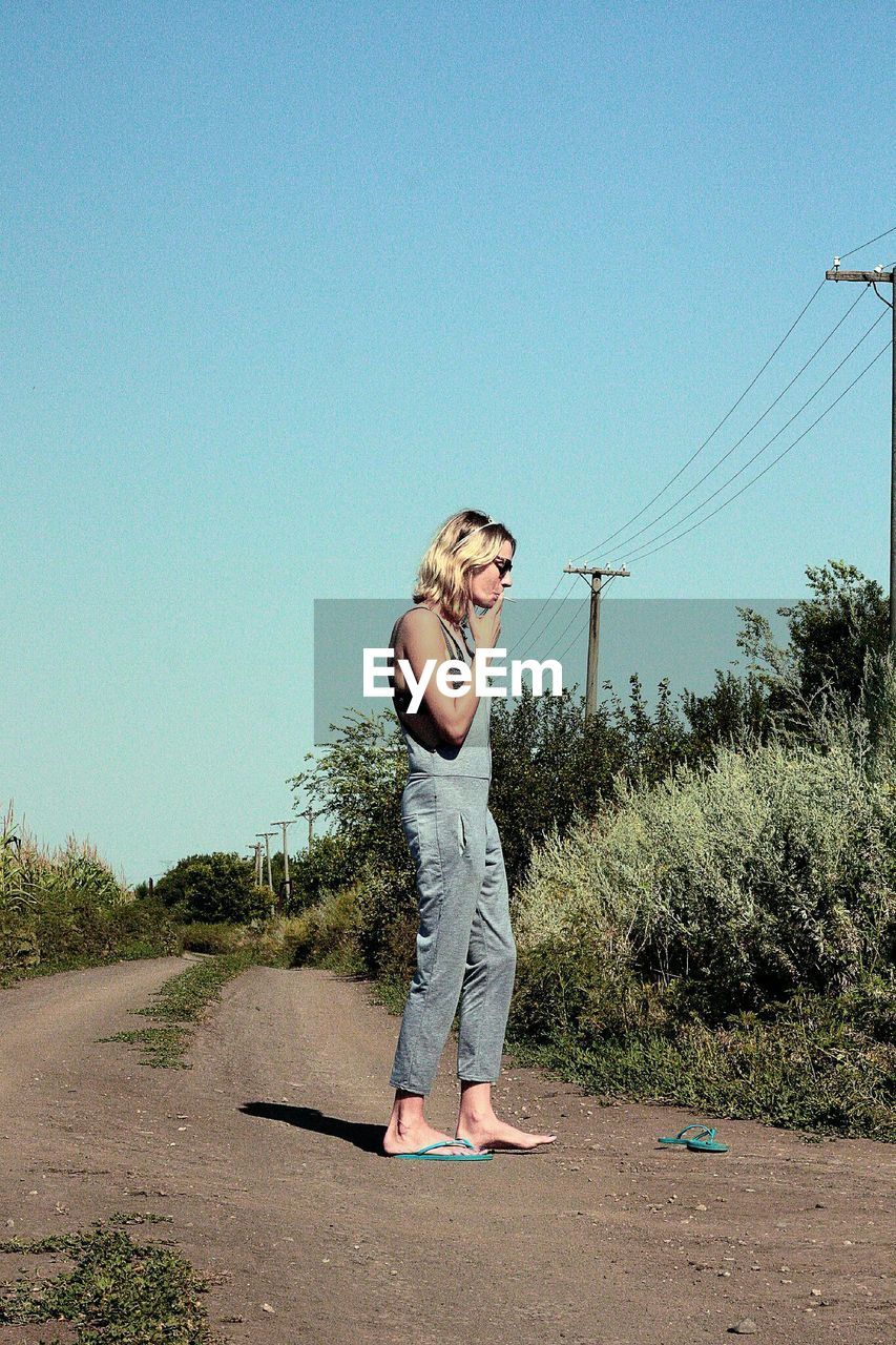 Side view of woman smoking cigarette on road against clear blue sky