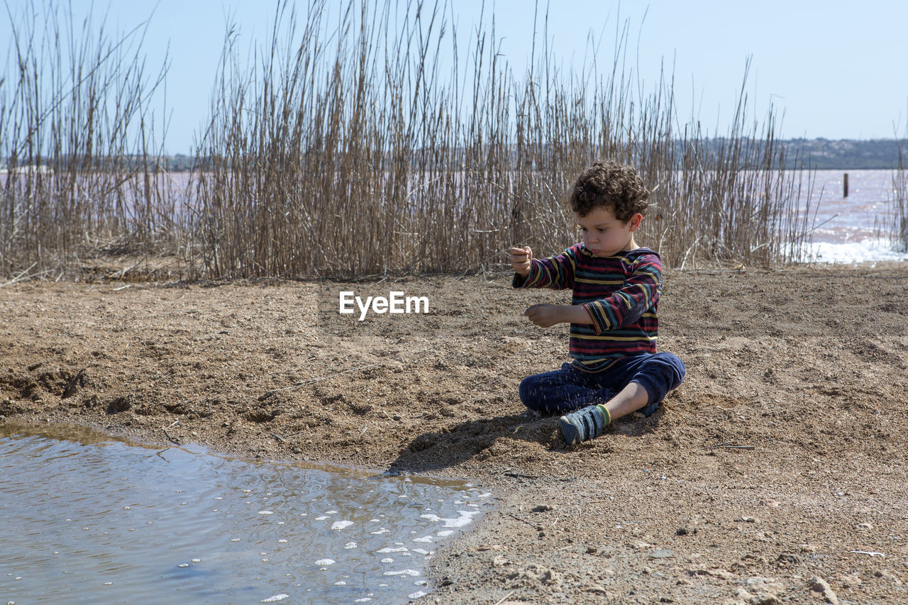 Small boy of 4-5 years playing with sand at the edge of pond on sunny day with colored striped shirt