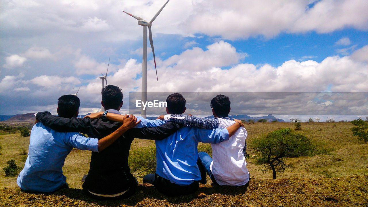 Rear view of friends sitting on field against windmills