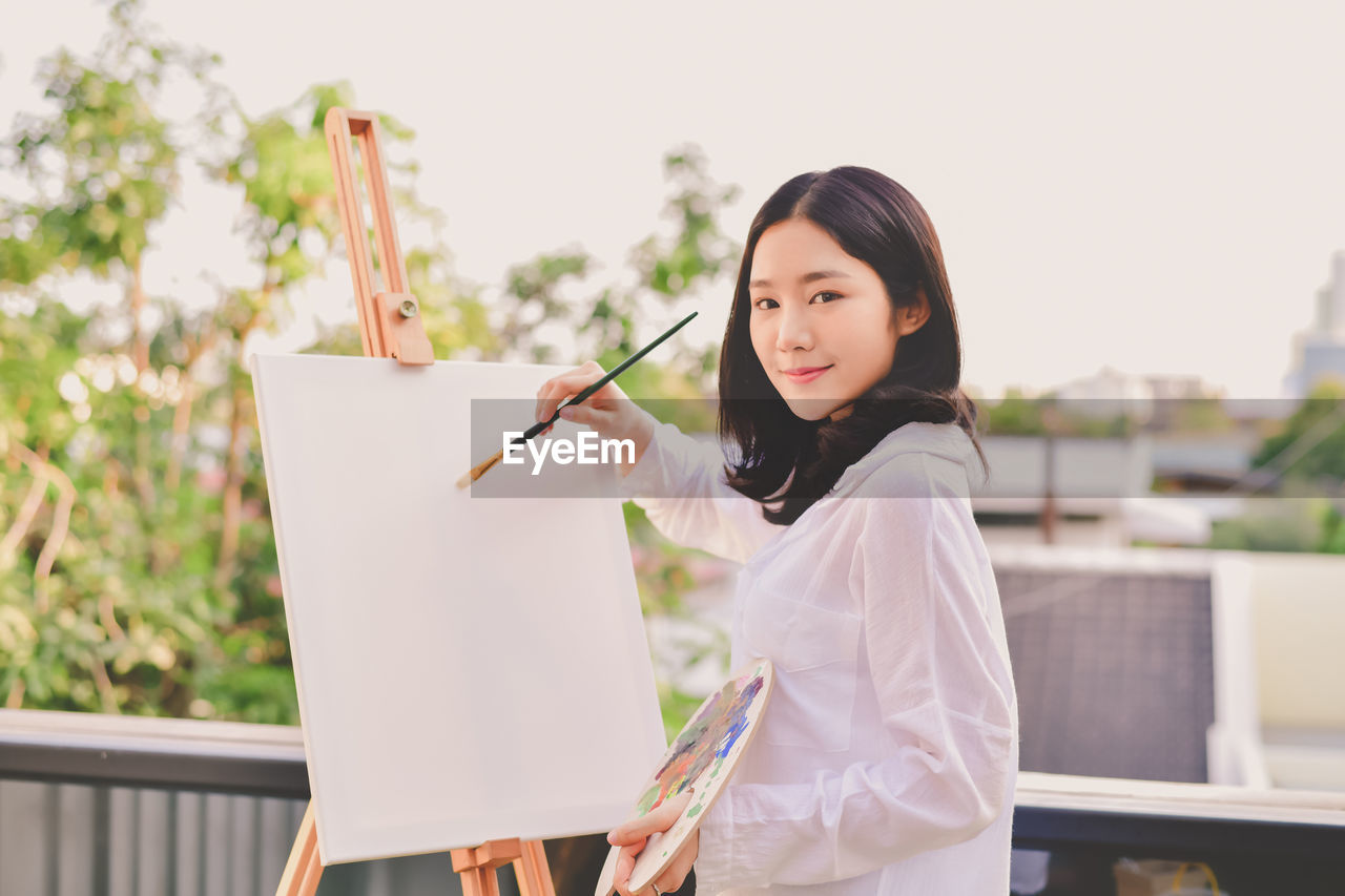 Portrait of woman painting on canvas against sky