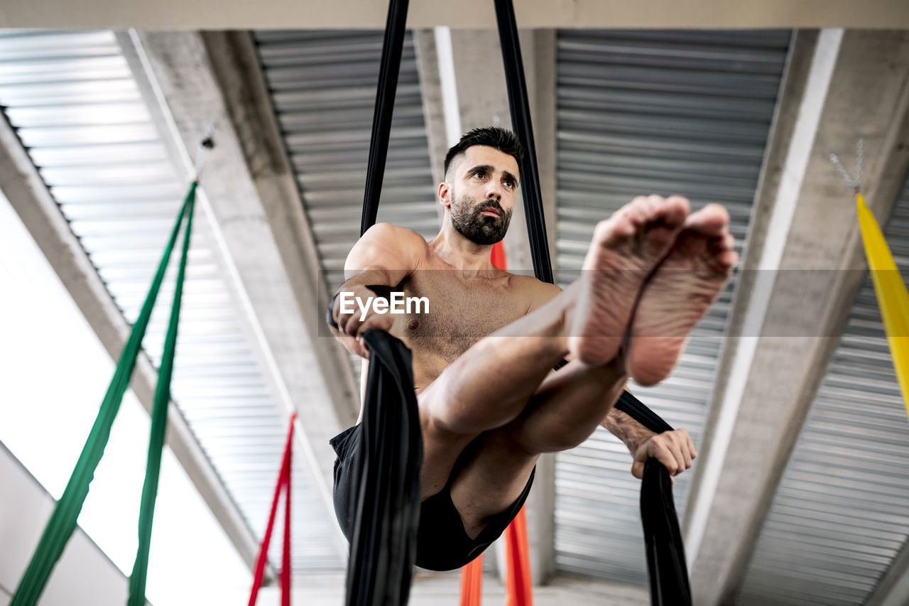 From below barefoot bearded man balancing on pieces of fabric and looking away against ceiling while practicing aerial dance in studio