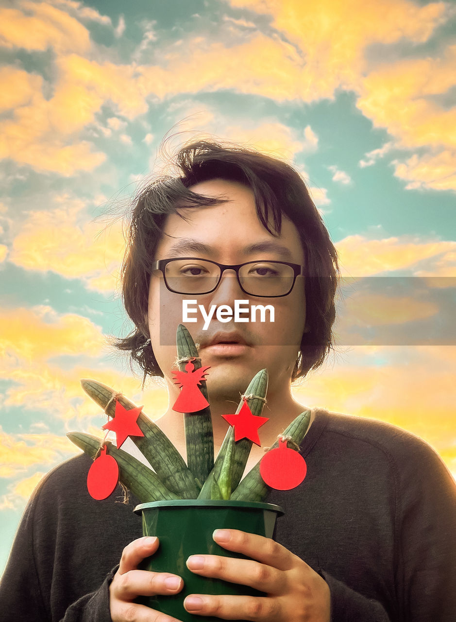 Portrait of young man holding potted succulent plant with christmas decorations against sunset sky