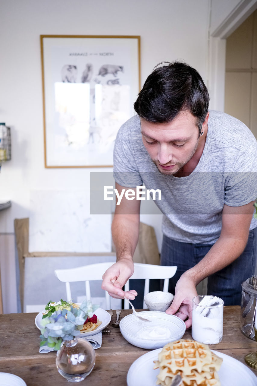 Man spreading yogurt in plate on wooden table against wall at home