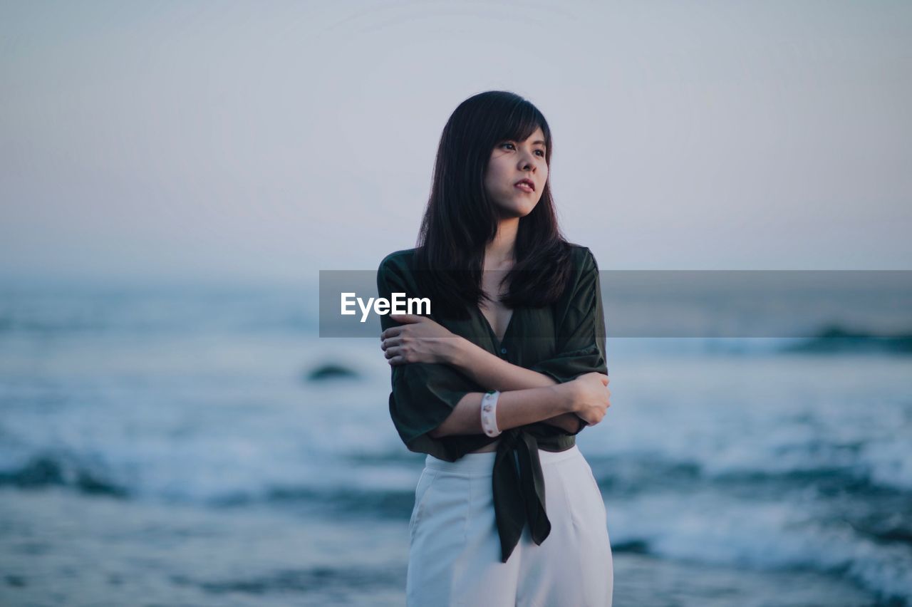 Thoughtful young woman hugging self while looking away at beach against sky