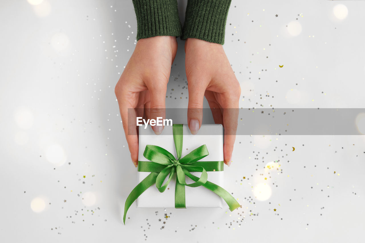 Hands holding xmas gift package in white wrapping paper with green ribbon on festive background