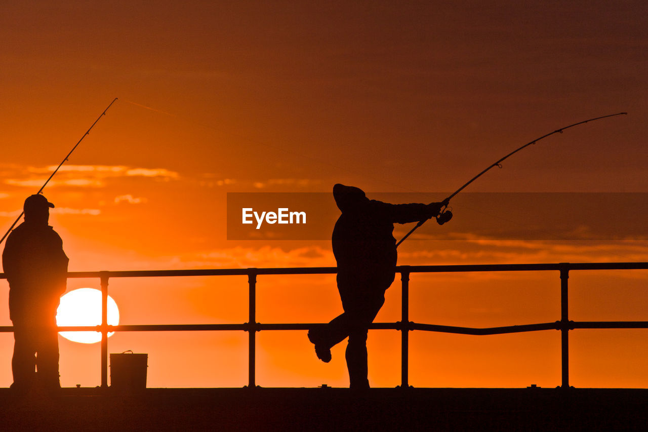 Silhouette men fishing by railing against sky during sunset