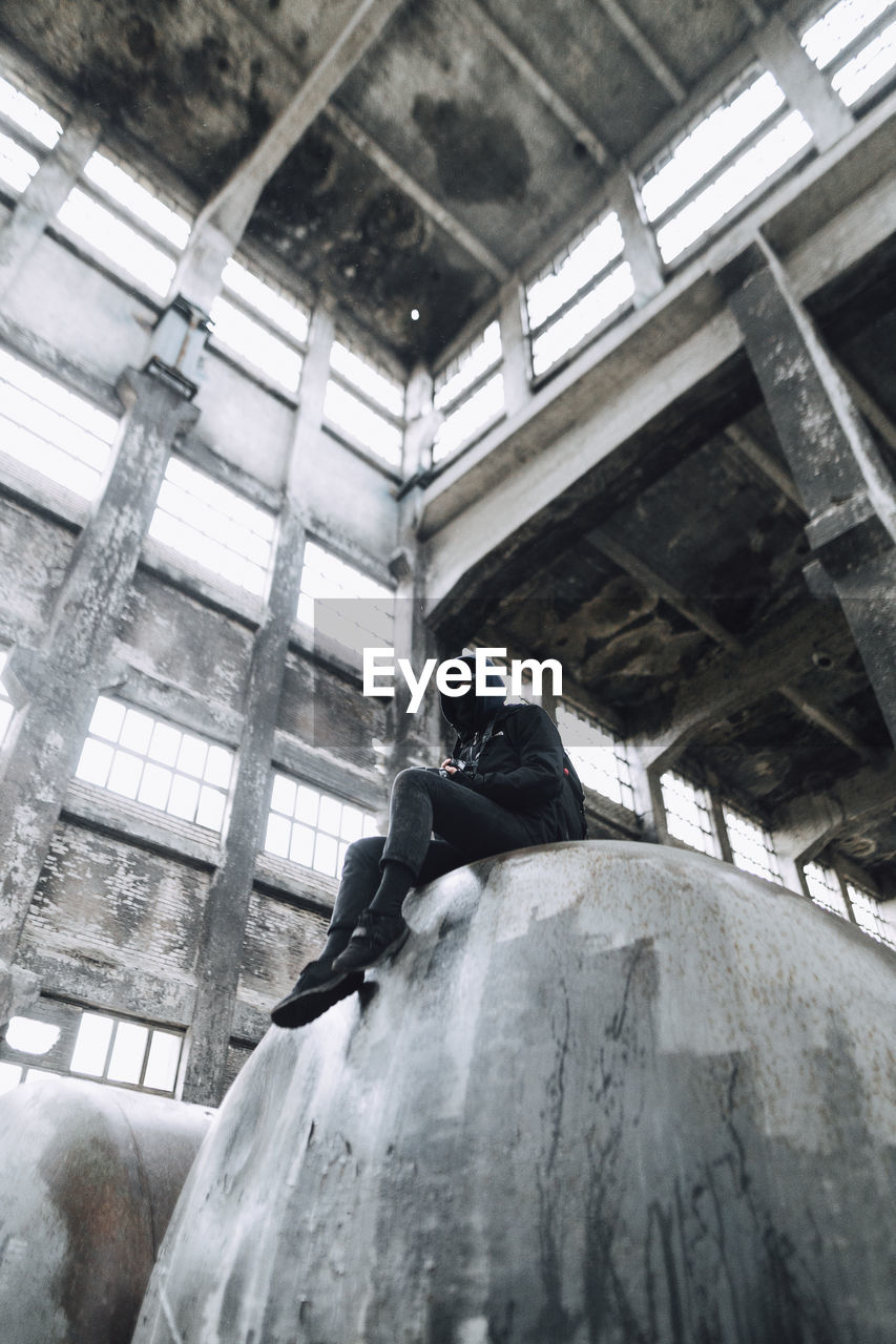 LOW ANGLE VIEW OF MAN SITTING ON ABANDONED BUILDING