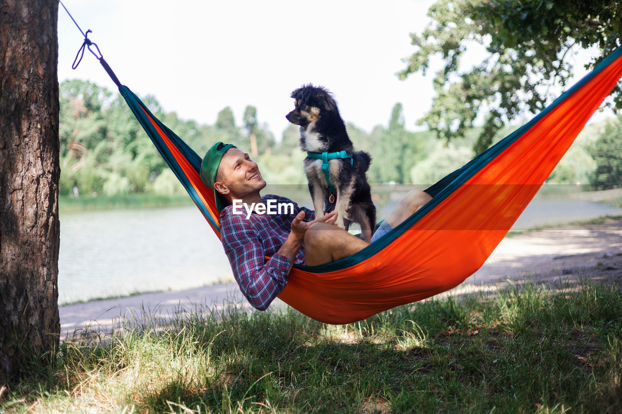 Man swinging with dog on hammock against trees in forest