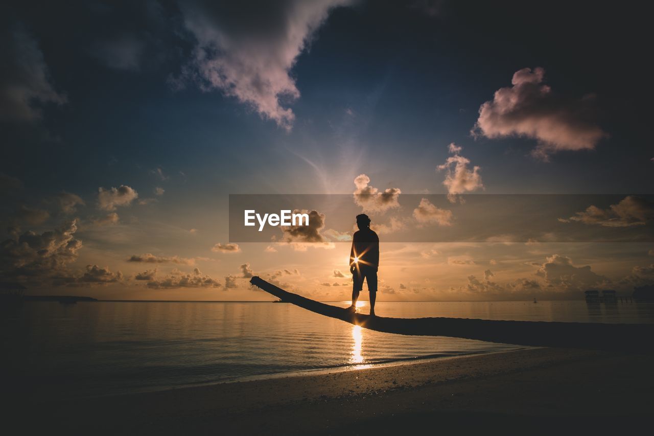 Silhouette man standing on tree trunk at beach against sky during sunset