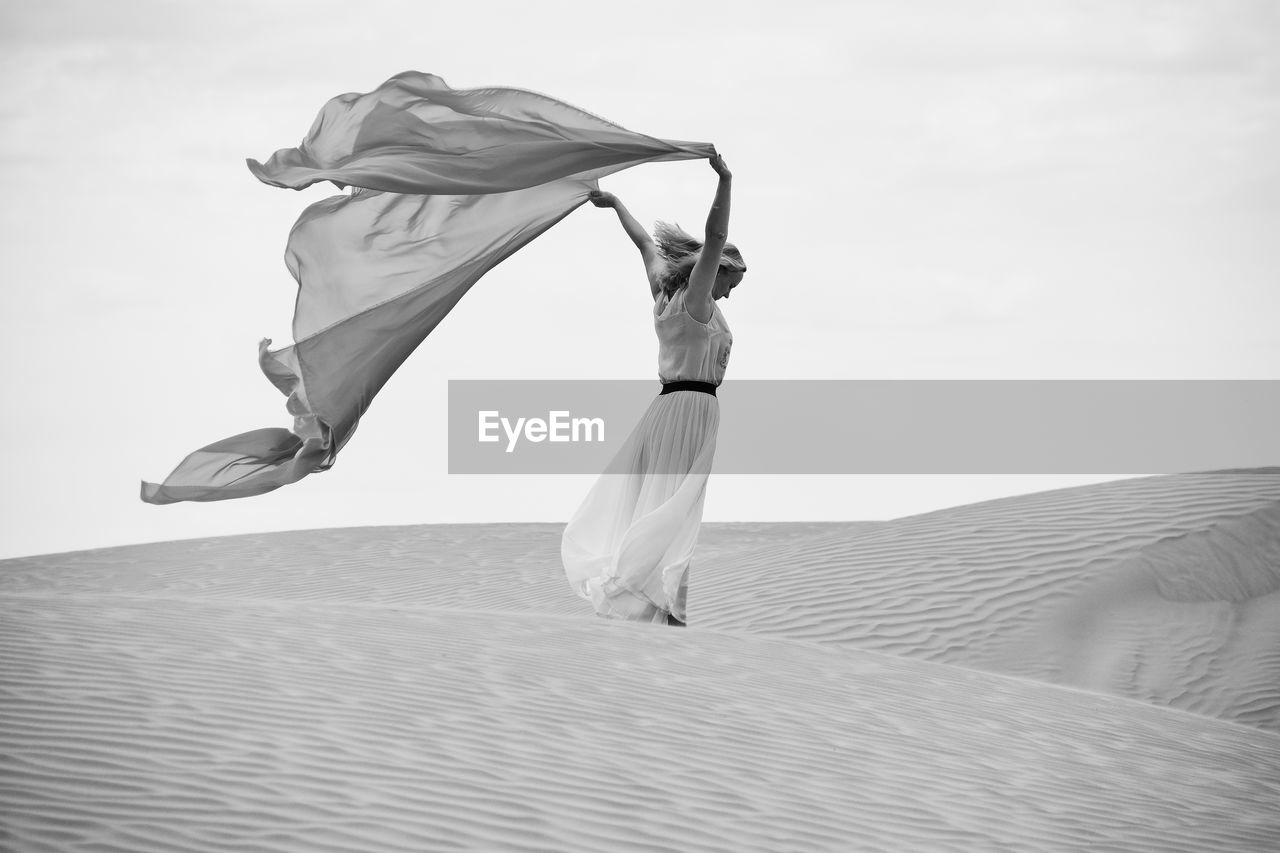 Woman with fabric standing on sand at beach against sky