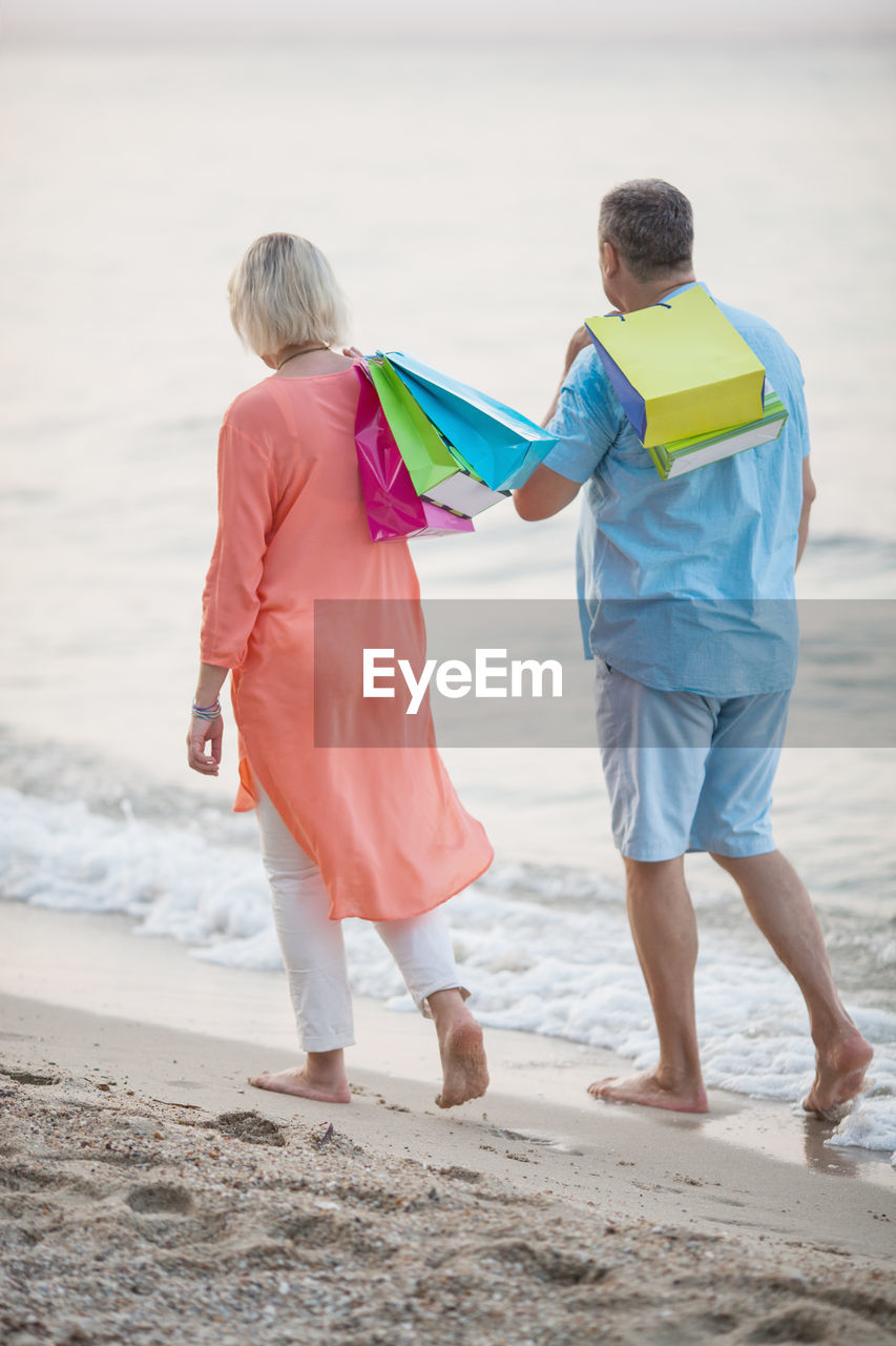 Rear view of mature couple with shopping bags walking on shore at beach