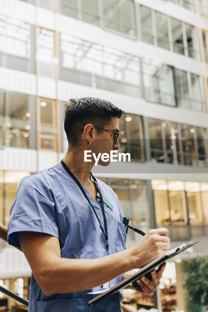 Mature male healthcare worker looking away while writing in clipboard at hospital