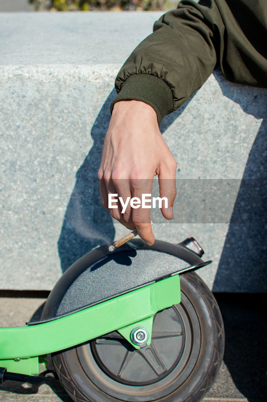 Cropped hand of man smoking joint with push scooter against railing