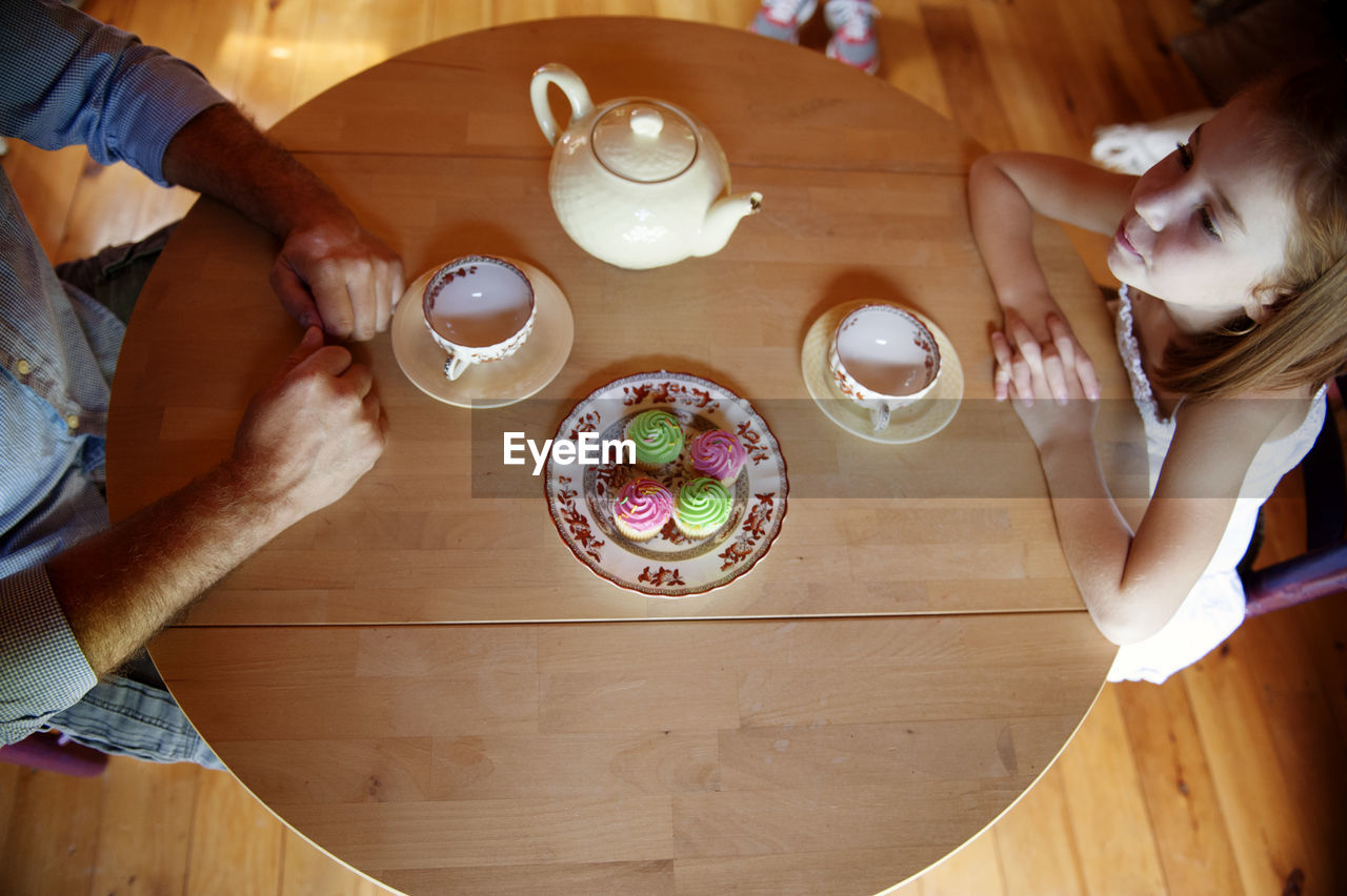 Overhead view of father and daughter sitting at table with cupcakes and tea