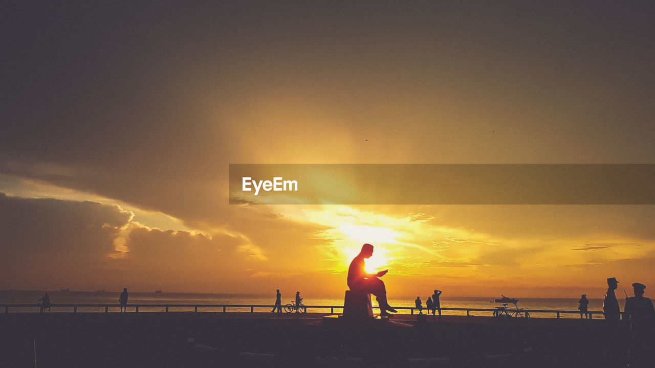 SILHOUETTE PEOPLE STANDING ON BEACH AGAINST ORANGE SKY