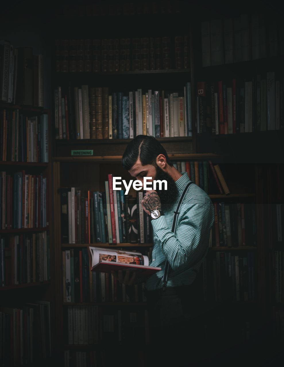 Man reading book while standing against shelves