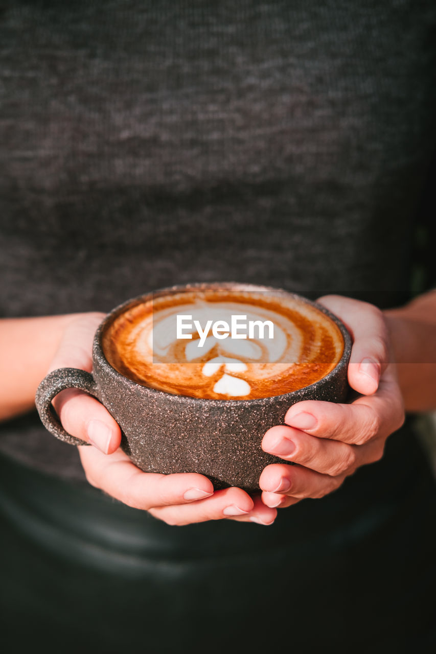 Woman in a black clothing holds coffee in a grey ceramic cup, vertical photo.