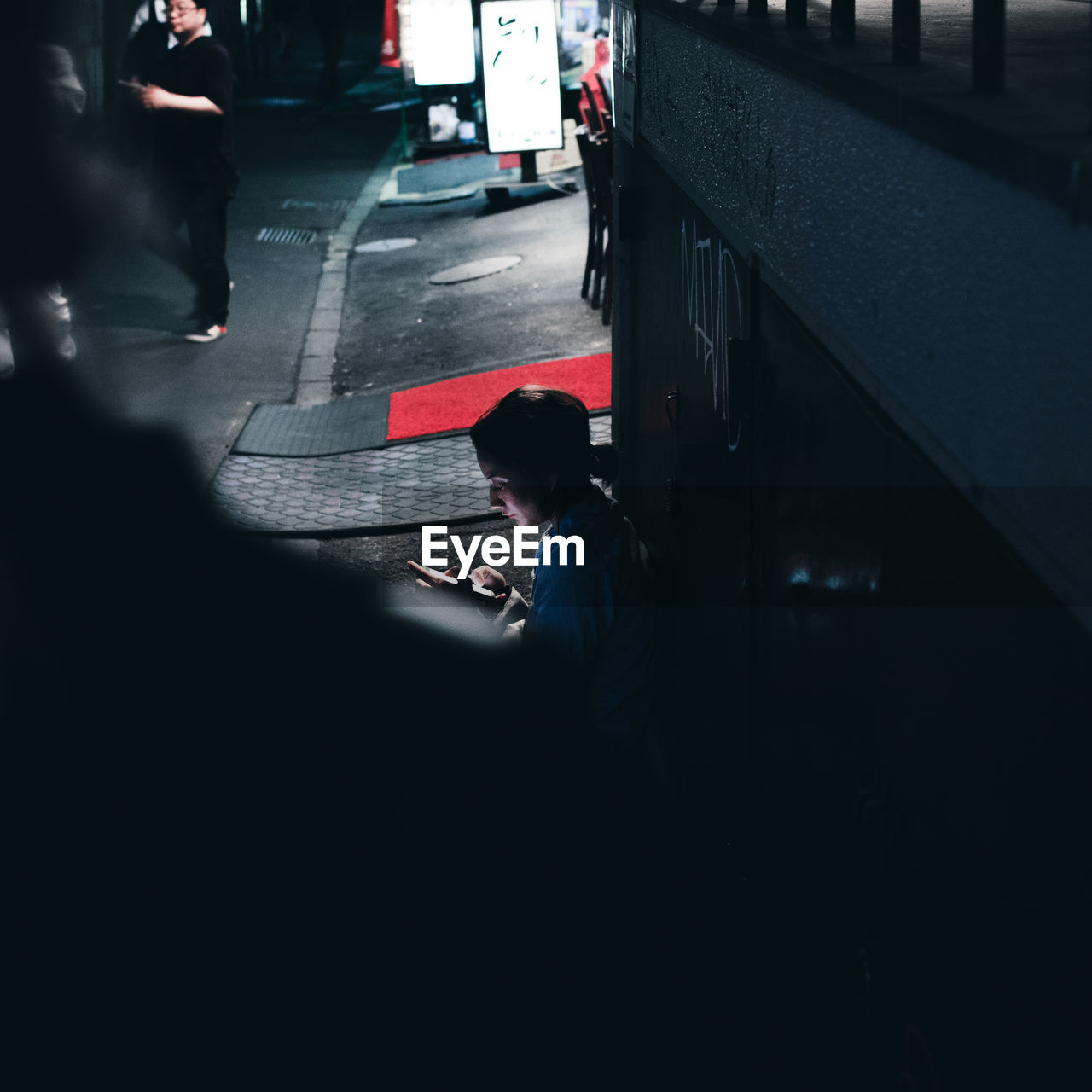 Side view of woman using mobile phone while sitting on sidewalk in city at night