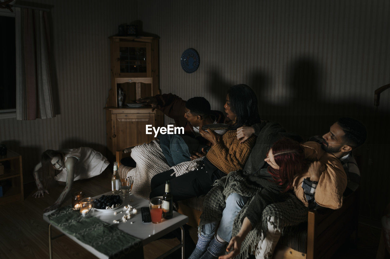 Frightened multiracial friends looking at spooky ghost while watching scary movie at cabin