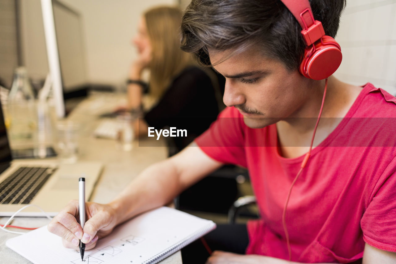 Young businessman wearing headphones while writing on book with colleague in background at creative office